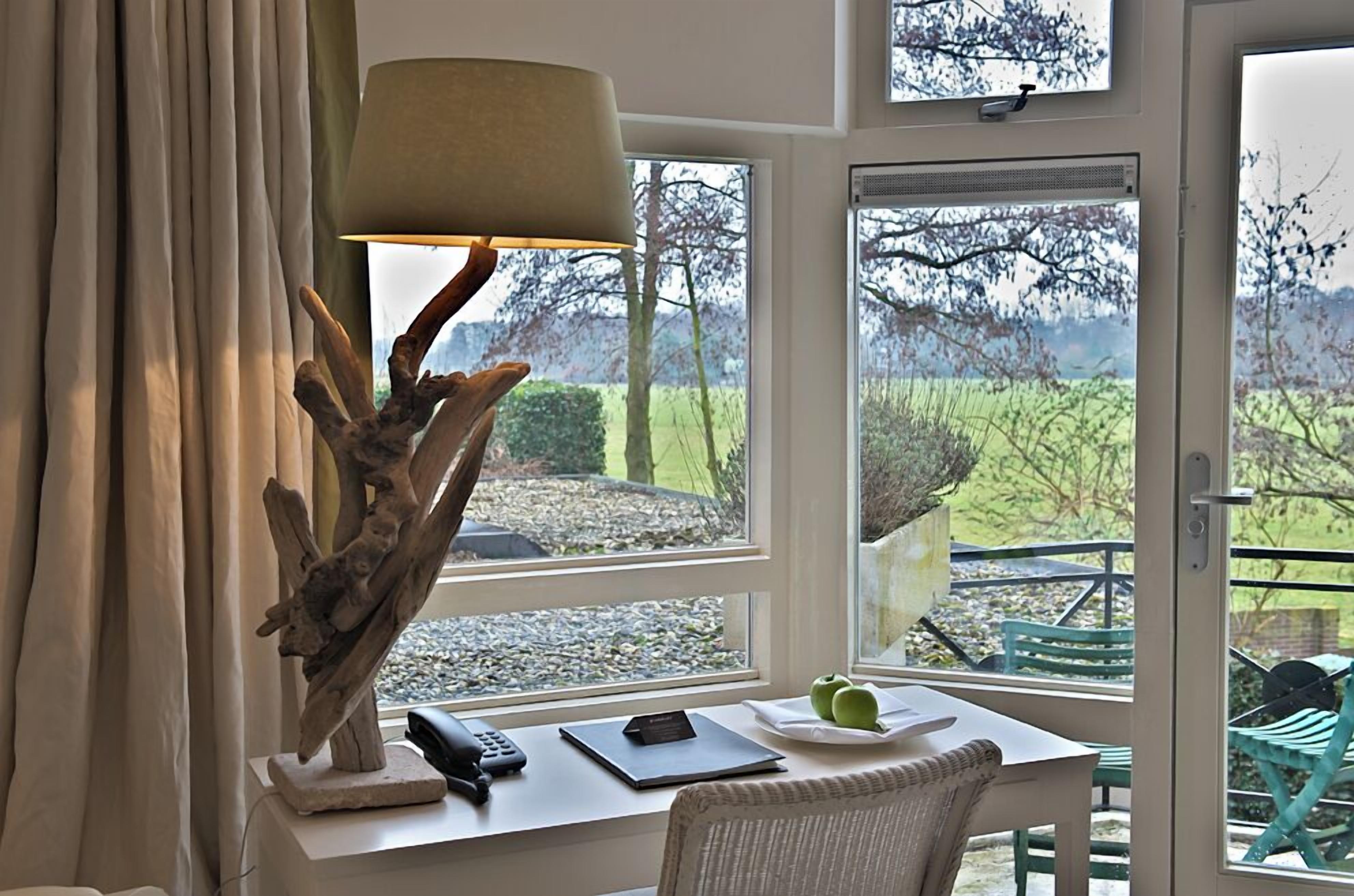A room with a desk, a lamp, a chair, and a view of the outdoors through large windows.