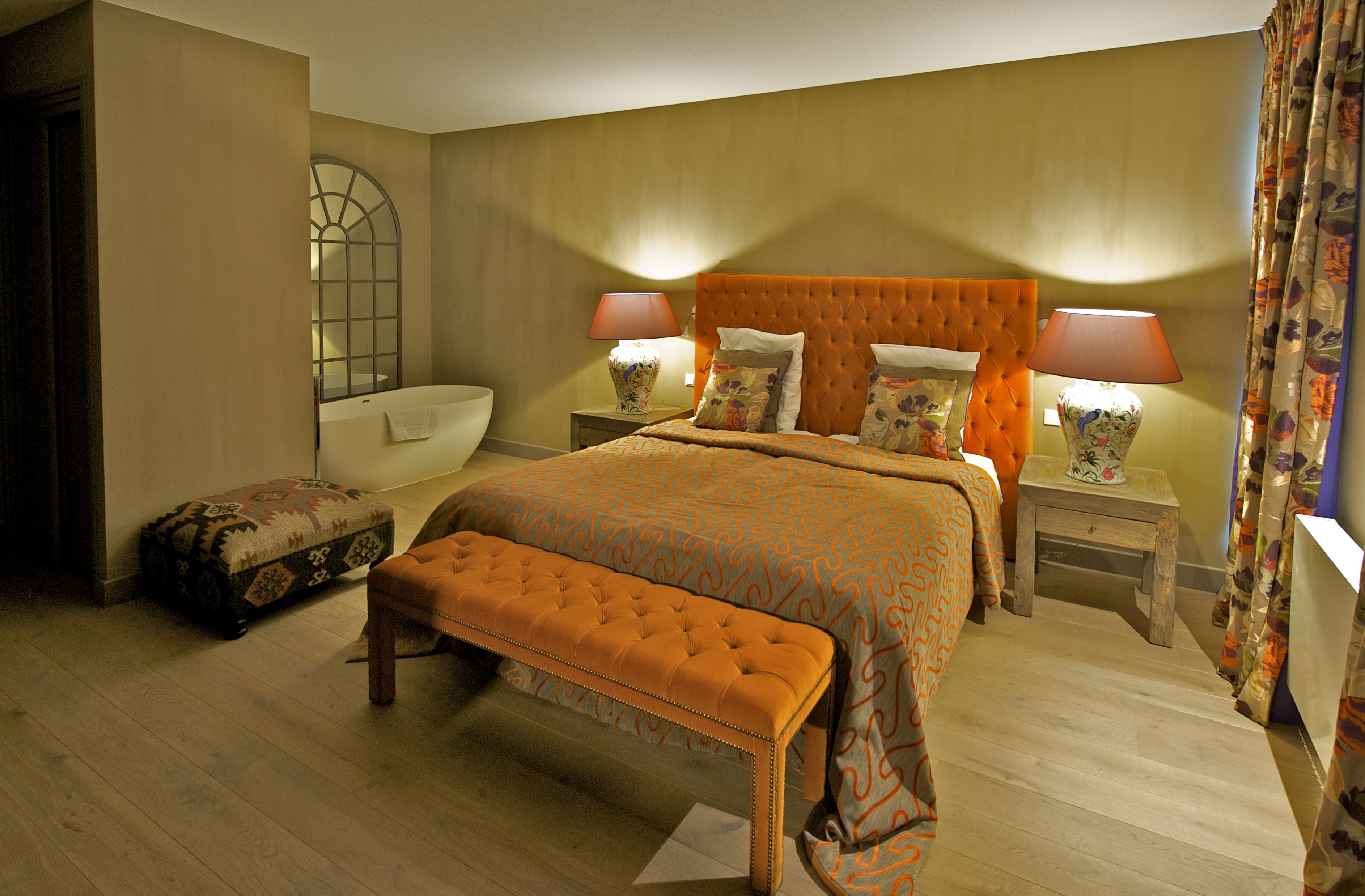 Een hotelkamer met een groot bed, een bankje, een nachtkastje met lamp, en een badkamer met spiegel en wastafel.