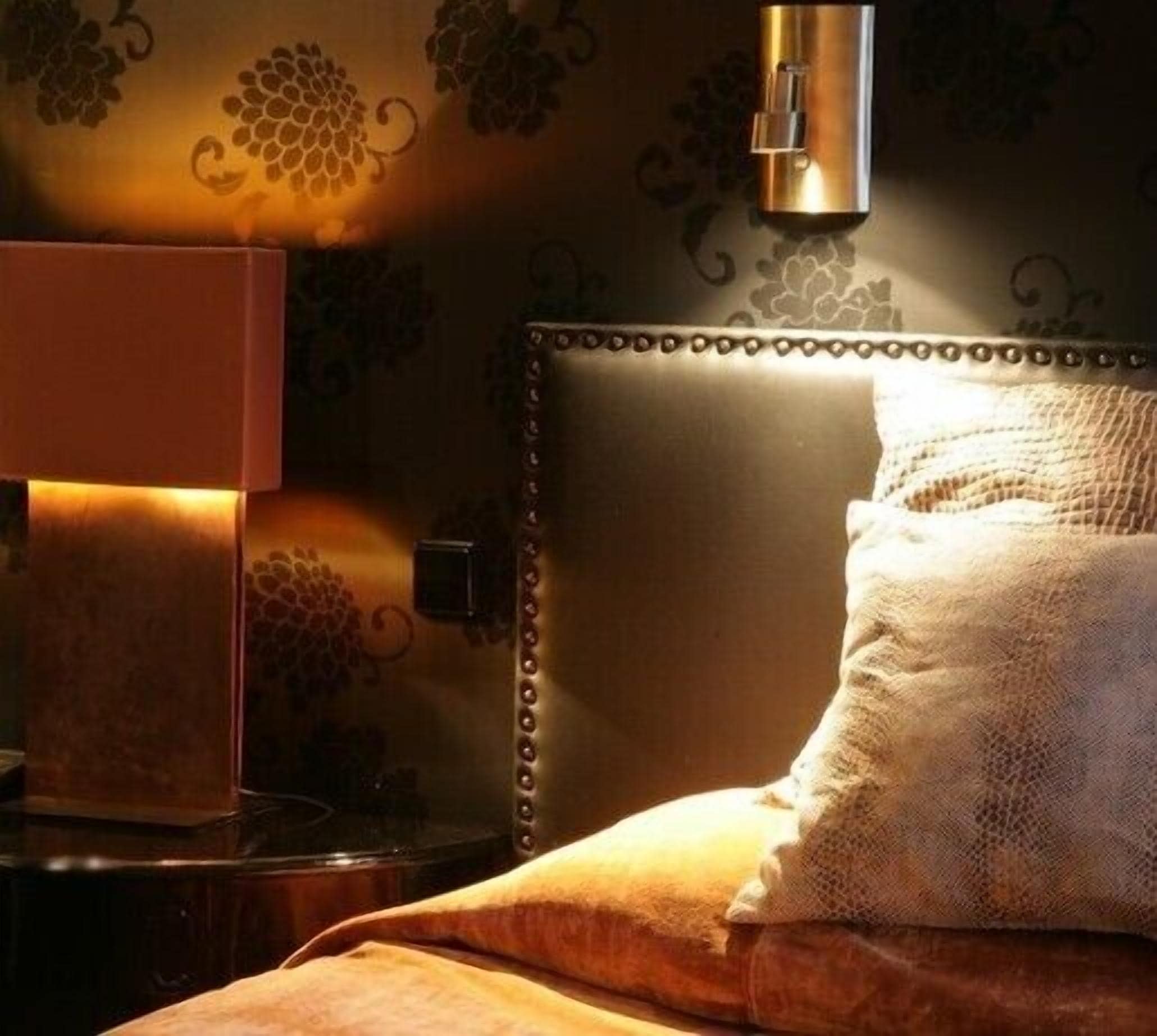Een keurig opgemaakt bed met een hoofdbord, een bedlampje en een muur met een druivenrankenpatroon.