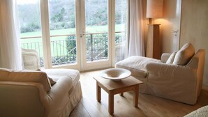 A room with a sofa, a table with a bowl, a lamp, and a door leading to a balcony with a view of greenery. - Restaurant Hotel Merlet  (Schoorl)