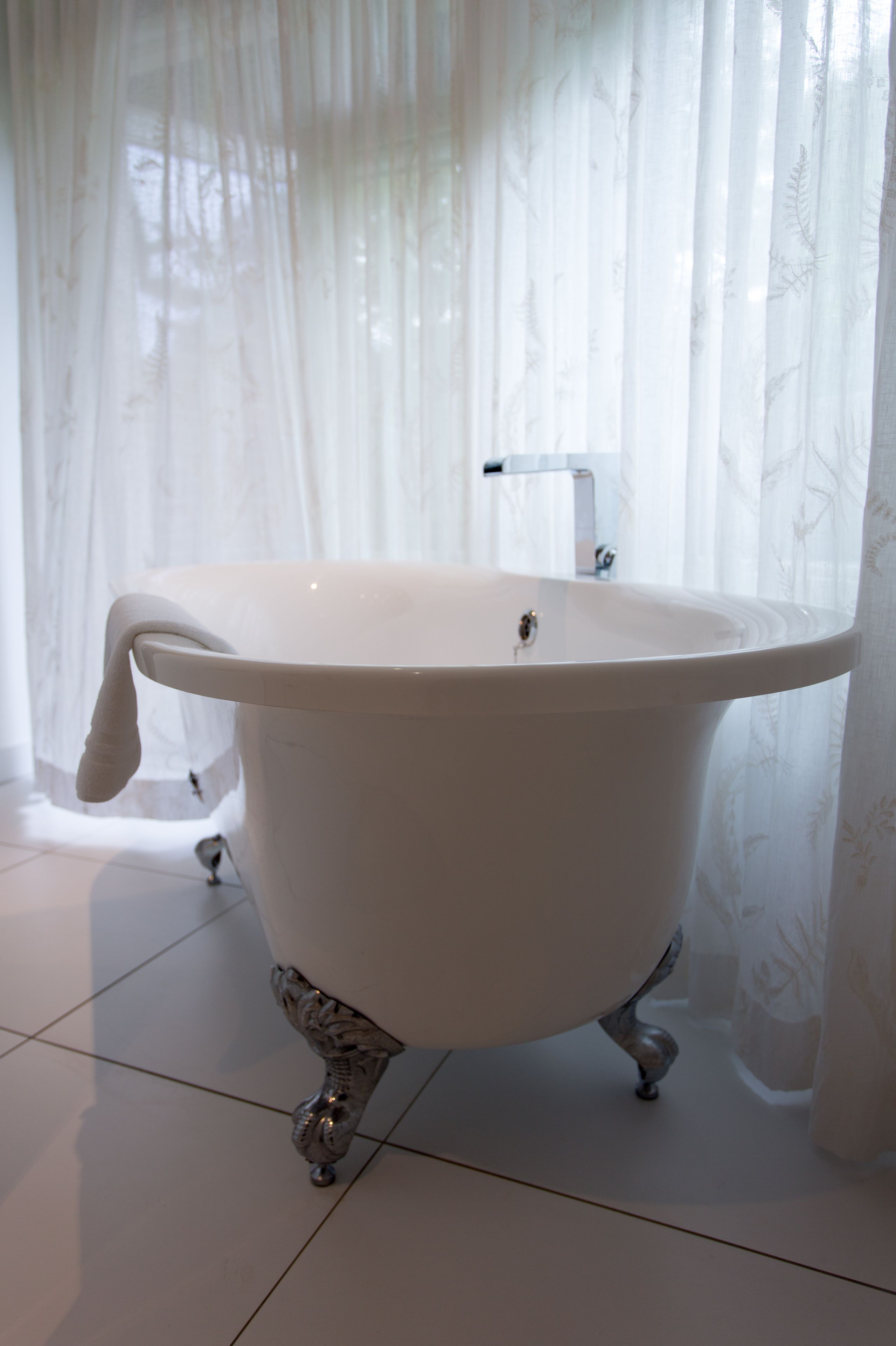 Eine weiße Badewanne auf Pfoten mit Wasserhahn, in einem Badezimmer mit weißen Fliesen und transparenten weißen Vorhängen.