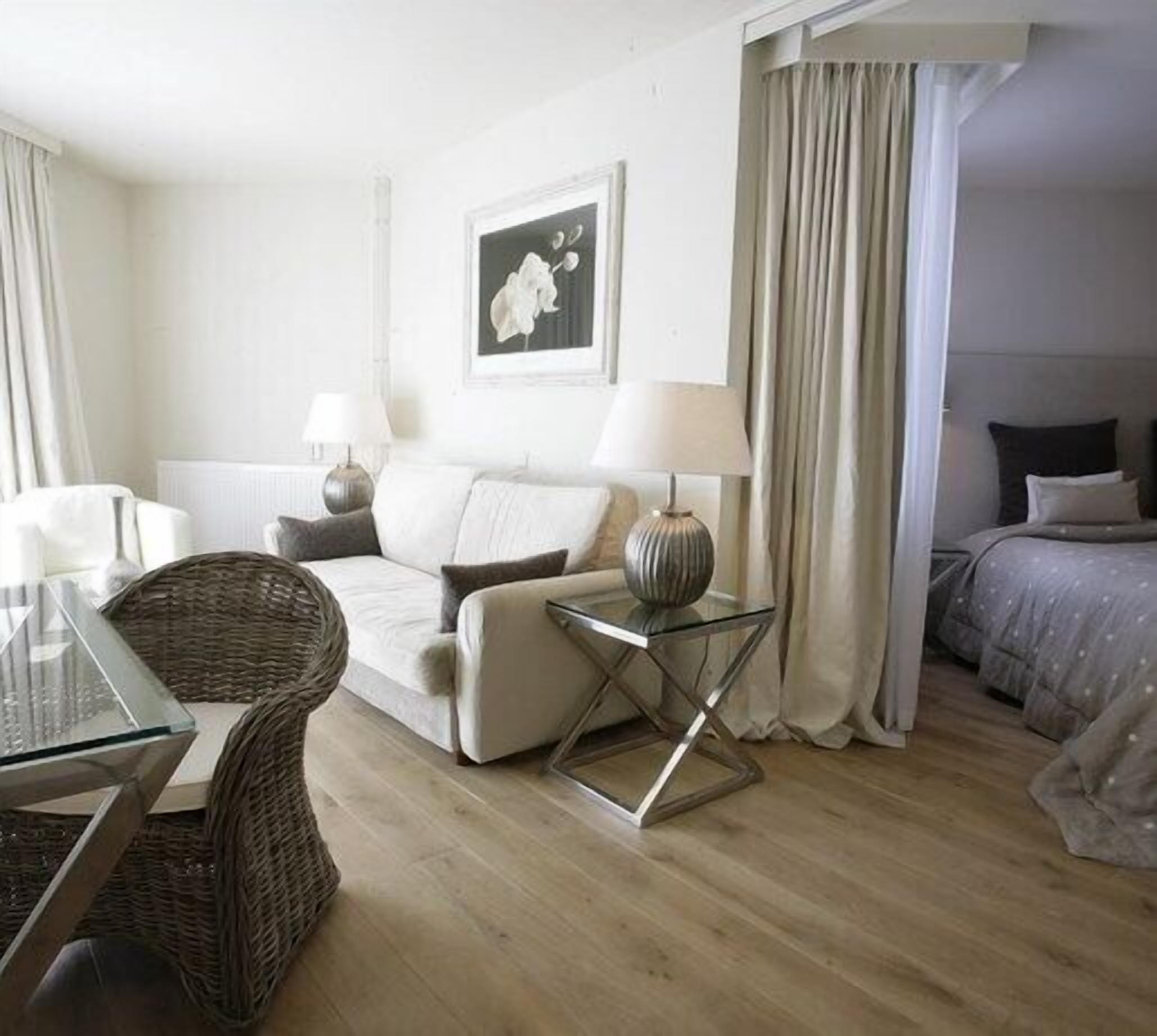 Une chambre d’hôtel avec un canapé, une table basse en verre, une table d’appoint avec une lampe, un lit avec une couette et un tableau encadré au mur.