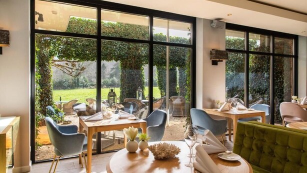 A dining area with large windows, a round table, and chairs. - Restaurant Hotel Merlet  (Schoorl)