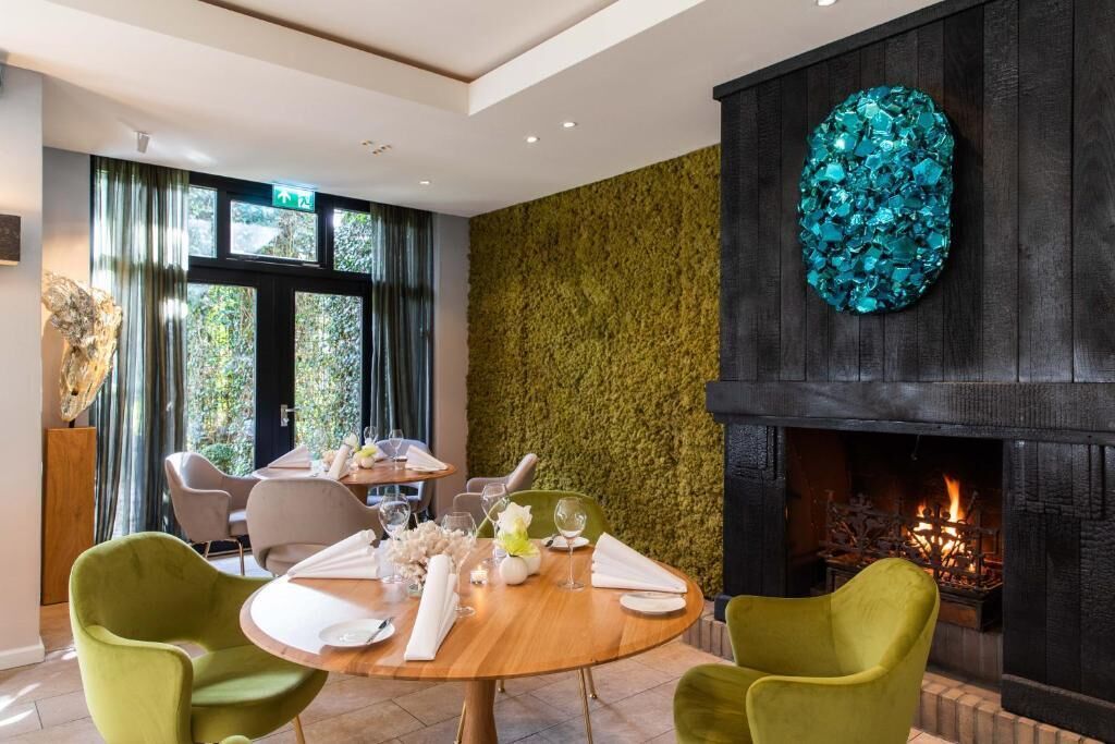 A dining area with a round wooden table set for two, surrounded by green upholstered chairs. A fireplace with a decorative teal mosaic is visible in the background.
