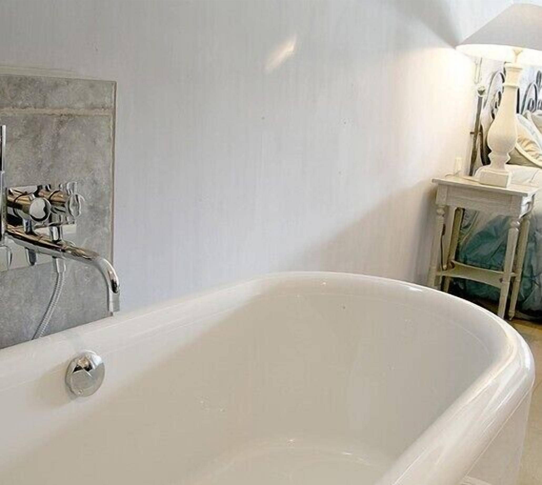 Un baño con una bañera blanca, una mesita con una lámpara y un revestimiento de azulejos.