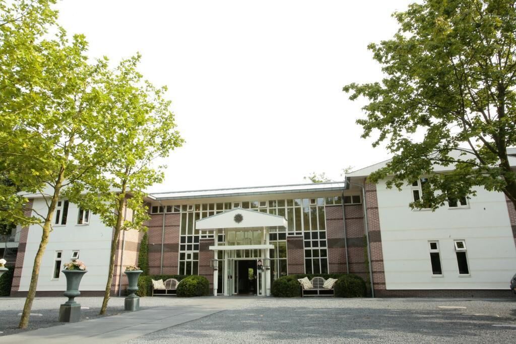 Un bâtiment doté d’une entrée centrale, bordée d’arbres et de jardinières, et une allée de gravier menant à l’entrée.