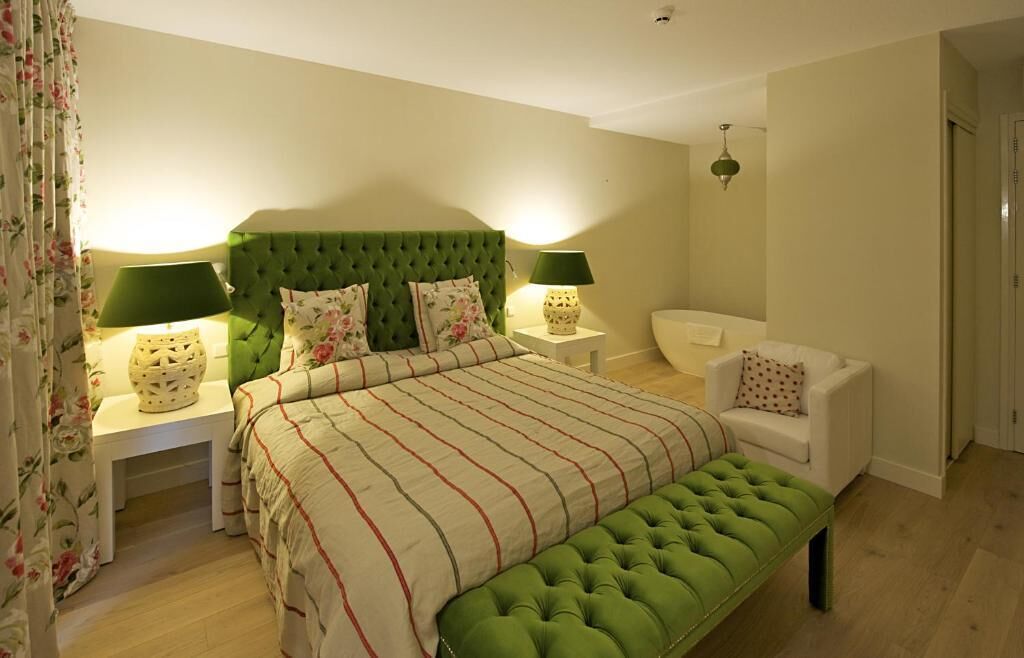 A bedroom with a green tufted headboard, a large bed with striped bedding, two bedside tables with lamps, a green upholstered bench, and a white armchair.