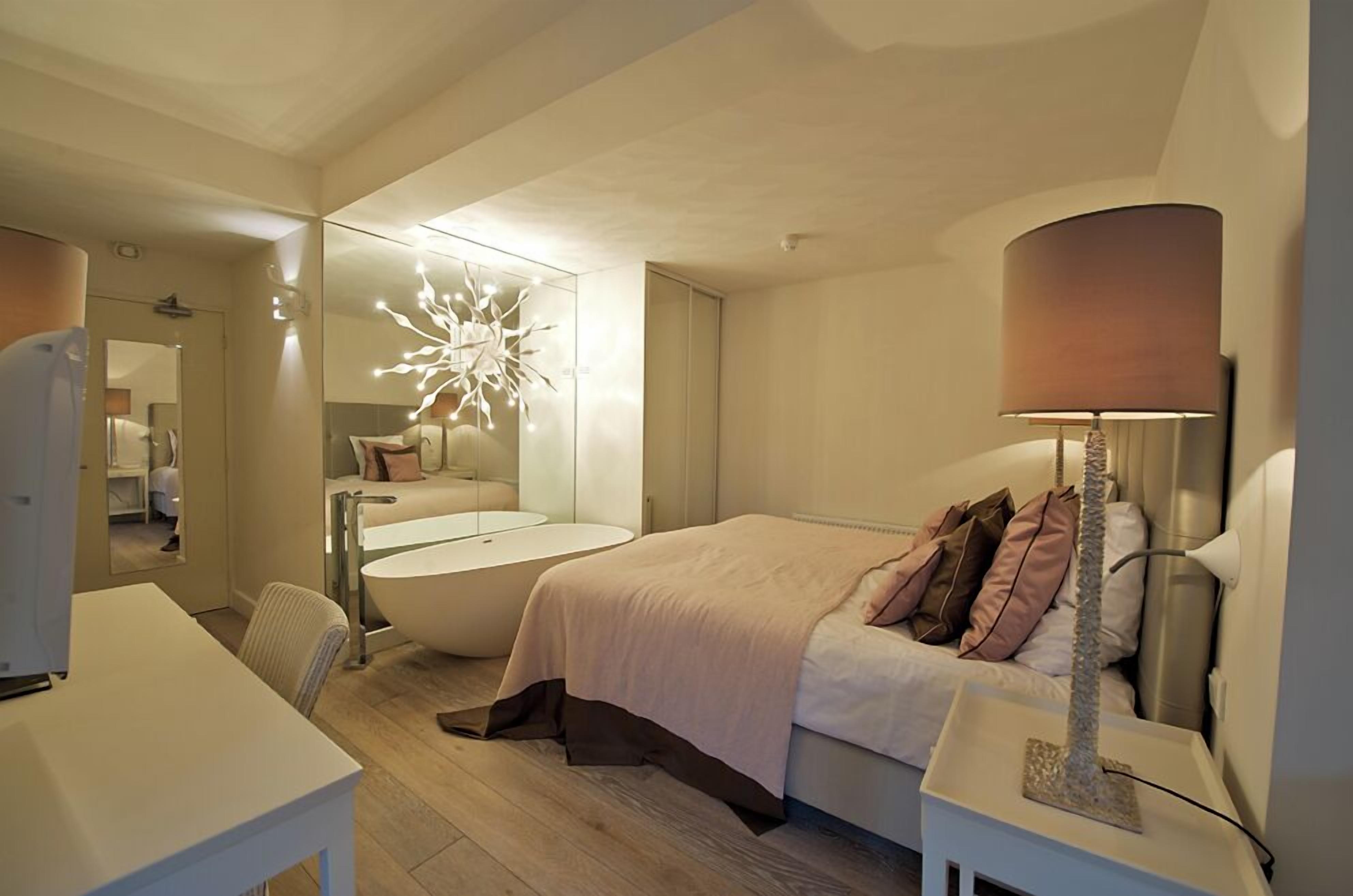 A modern hotel room with a large bed, a sitting area with a chair, a desk, and a bathtub.