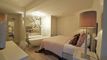 A modern hotel room with a large bed, a sitting area with a chair, a desk, and a bathtub.