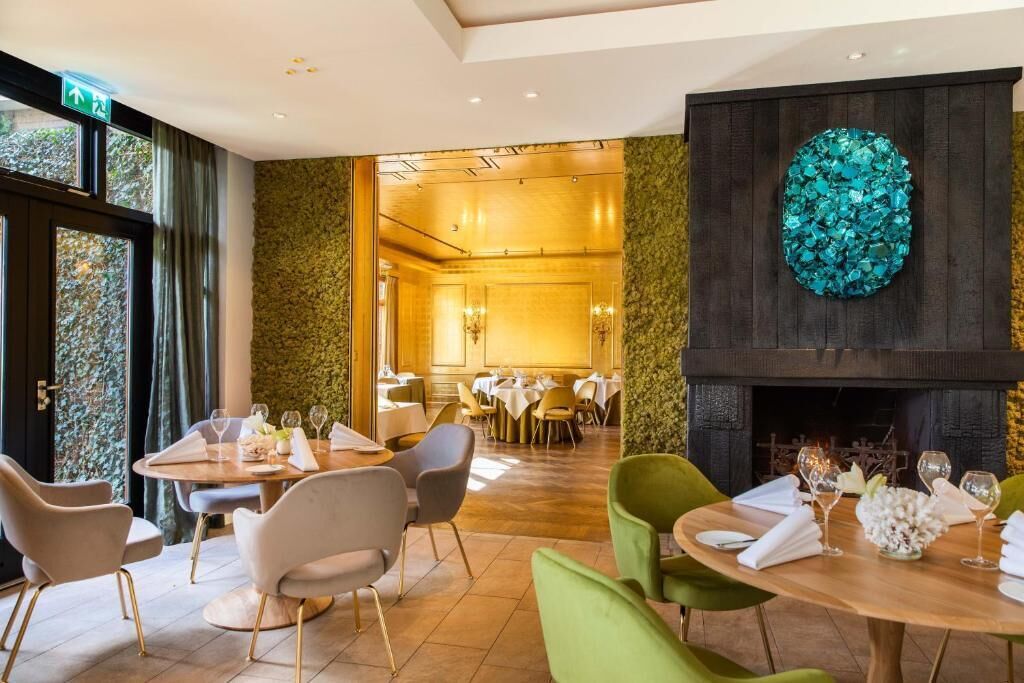 A restaurant with round tables and chairs, a fireplace, and a green wall with a decorative piece.