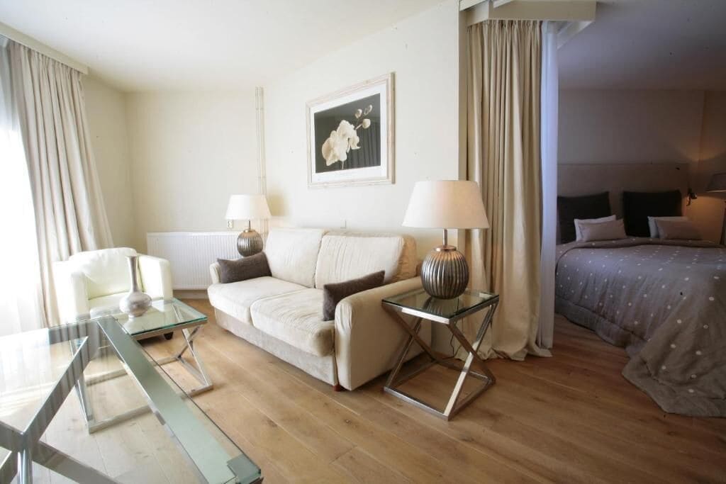 Une chambre d’hôtel avec un canapé, une table basse en verre, une lampe, un lit avec une couette grise et un tableau encadré au mur.