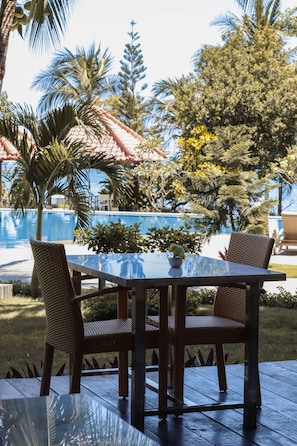 A glass-top table with wicker chairs, a potted plant, and a view of a swimming pool and palm trees. - LAPRIMA Hotel Flores (Labuan Bajo)
