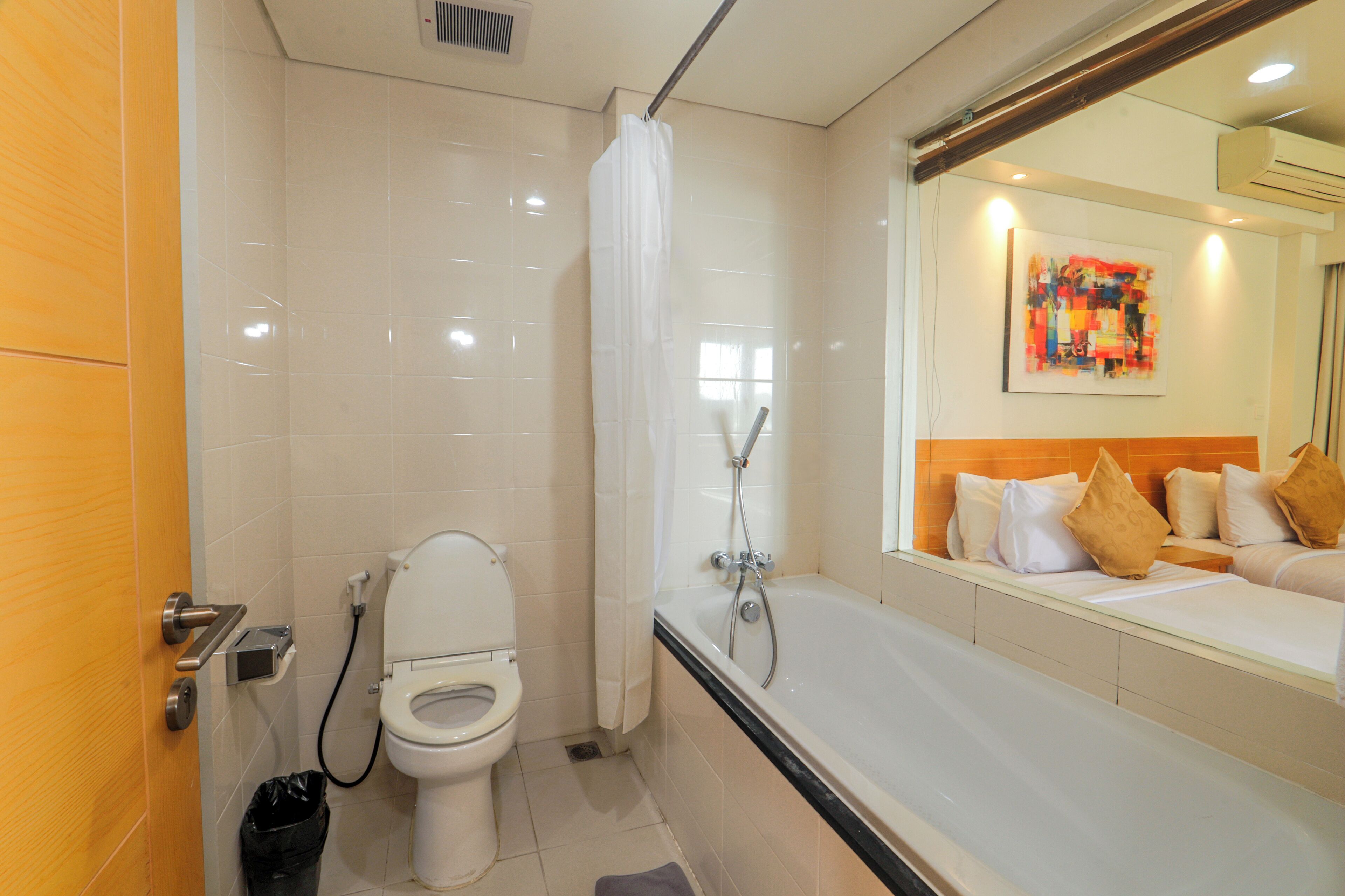 A modern bathroom with a toilet, shower, and a large mirror reflecting a bed with pillows.