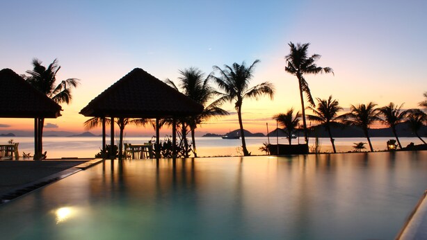 A poolside view with thatched-roof structures and palm trees at sunset. - LAPRIMA Hotel Flores (Labuan Bajo)