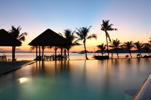 A poolside view with thatched-roof structures and palm trees at sunset.