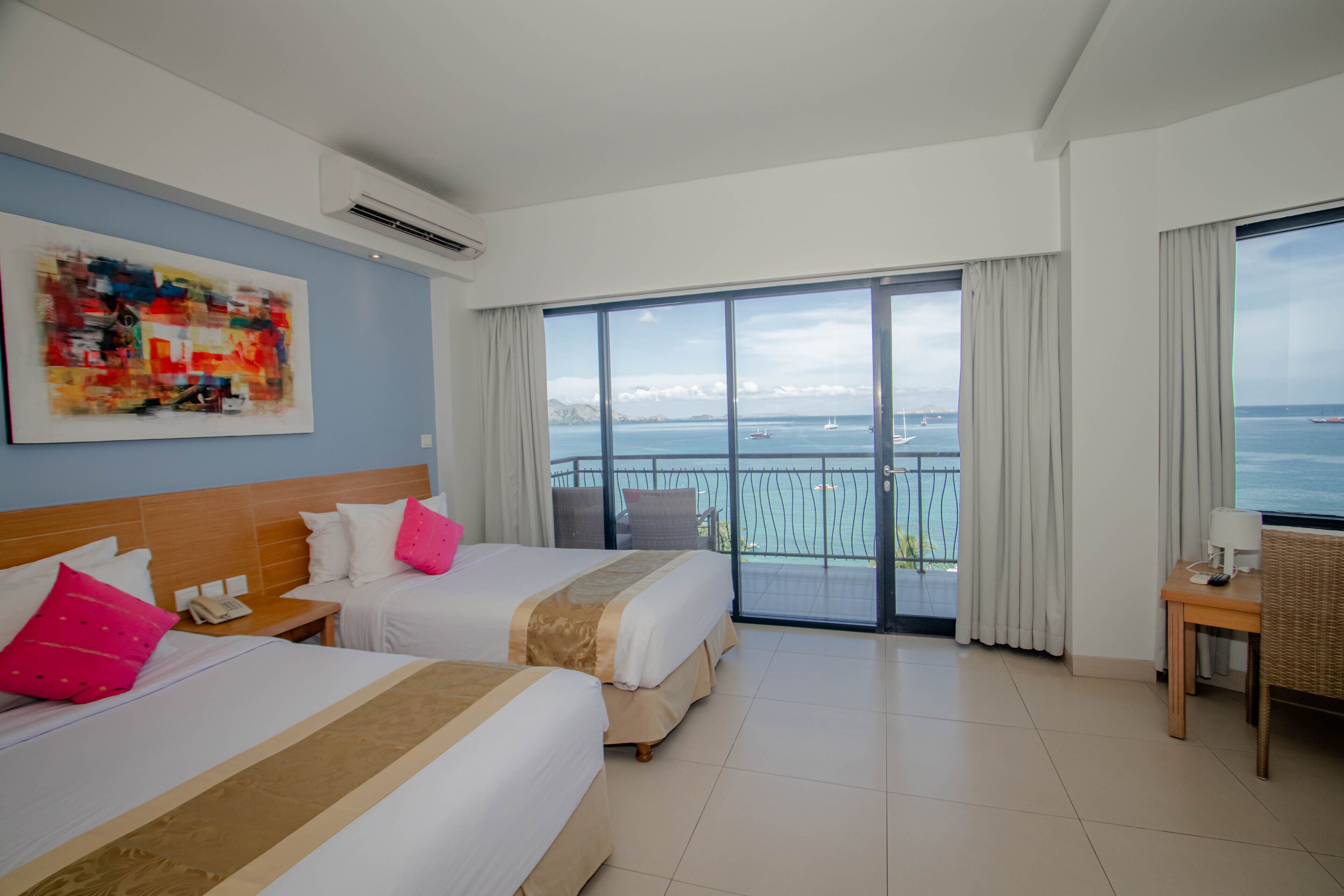 A hotel room with two beds, a large window with a view of the sea, a painting on the wall, and a small table with a lamp.
