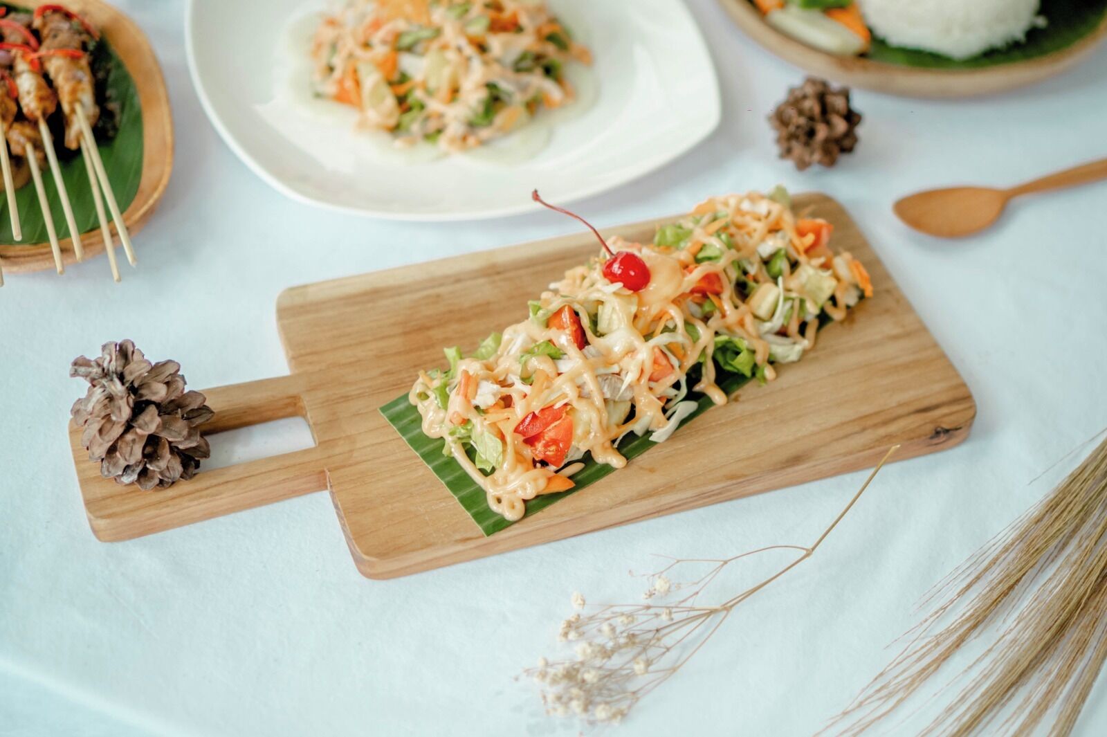 A wooden serving board with a salad and a cherry on top, placed on a table with a pine cone and dried flowers.
