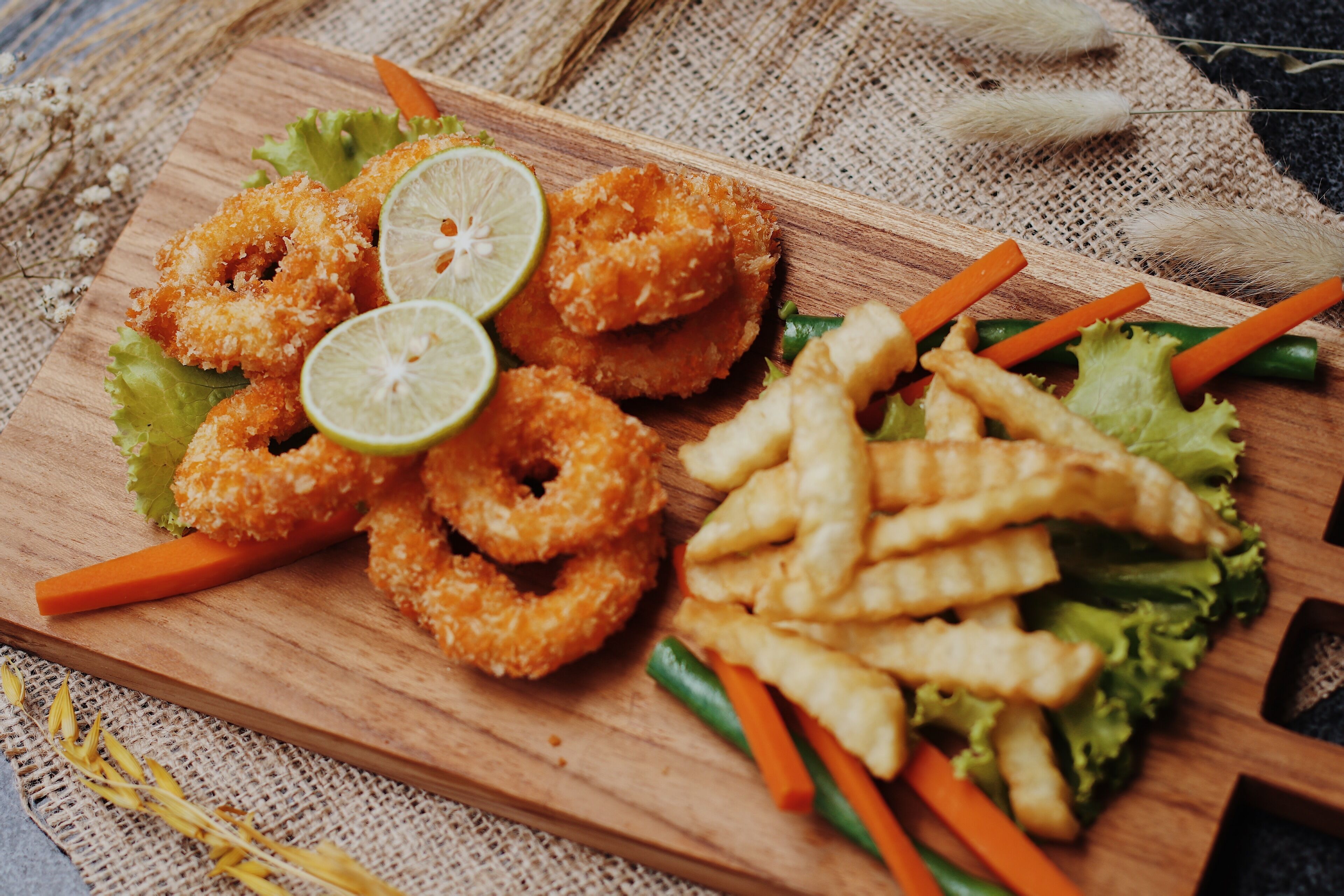 A wooden board with fried food items, including onion rings and fries, garnished with carrots and lettuce.