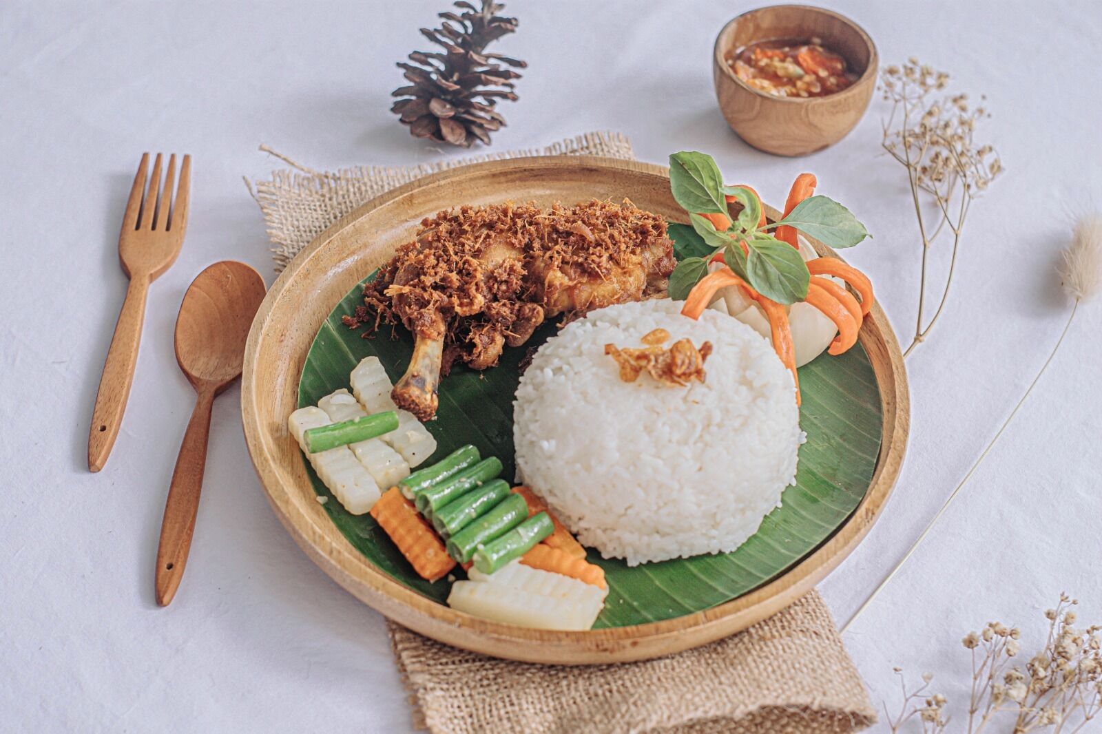 Un plato de comida con arroz, verduras y pollo frito sobre una hoja de plátano, acompañado de utensilios de madera y un pequeño recipiente con salsa.
