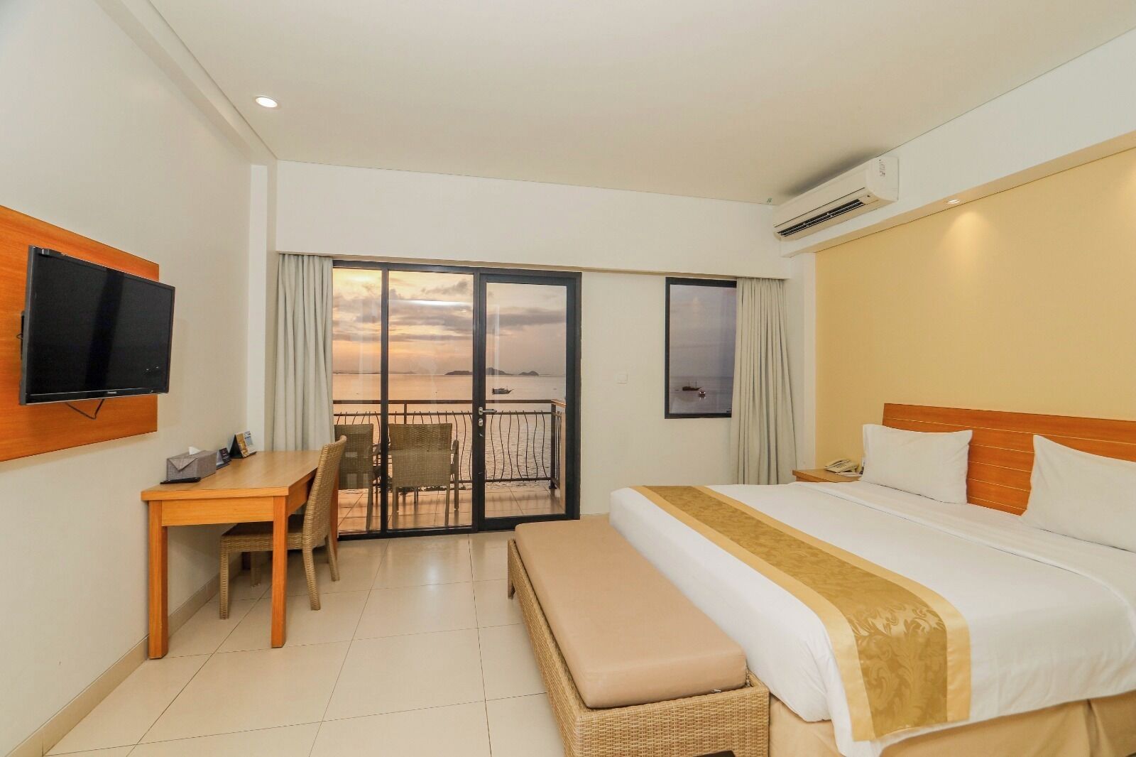 A hotel room with a bed, a desk, a chair, a television, and a balcony with a view of the sea.