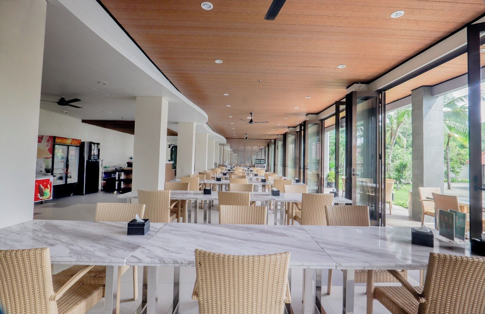 A spacious indoor dining area with marble tables and wicker chairs, large windows offering views of greenery, and a ceiling with recessed lighting.