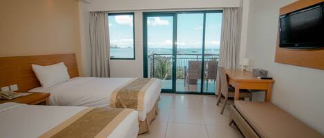 A hotel room with two beds, a desk, a TV, and a balcony with a view of the sea.