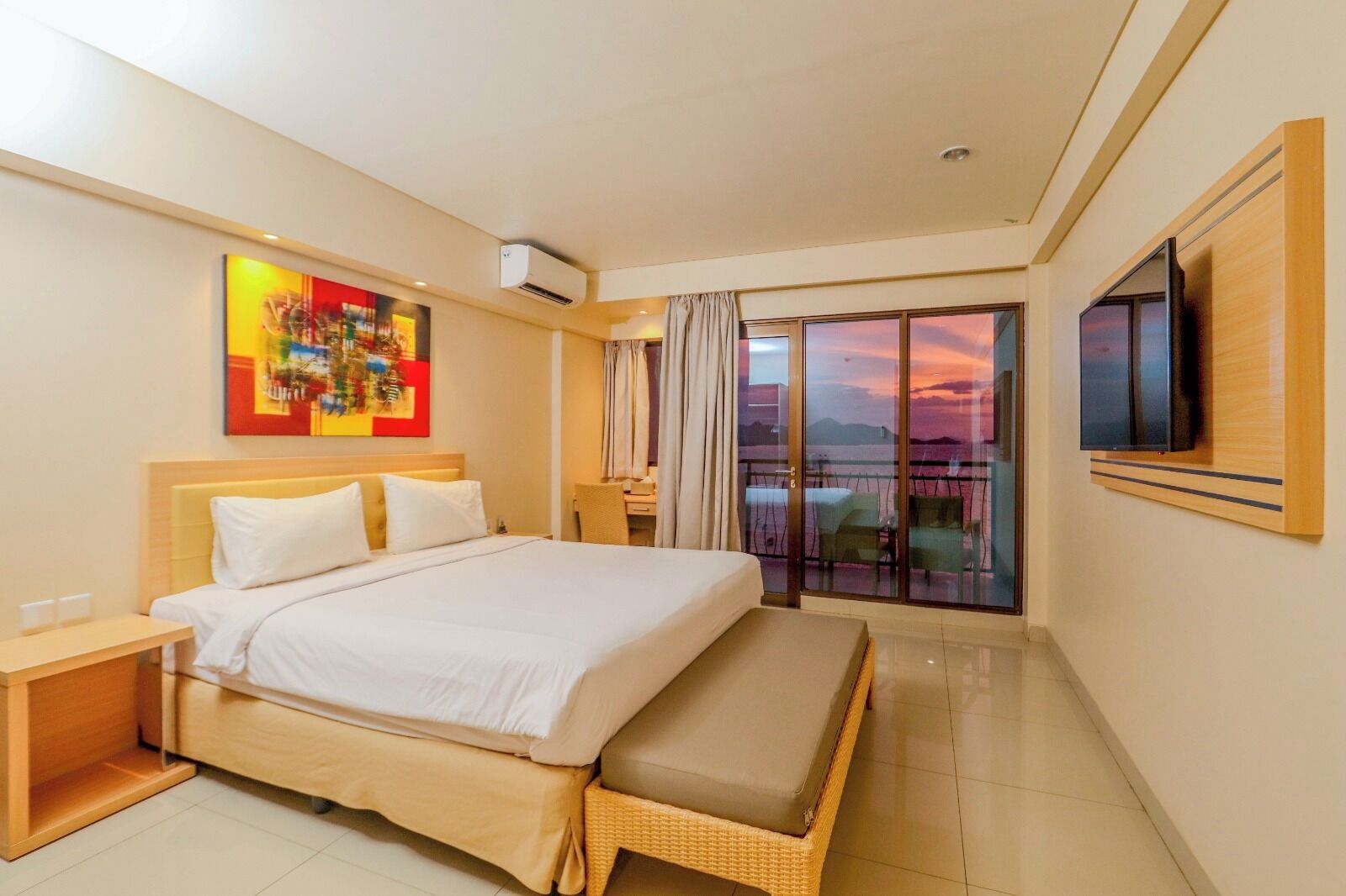 Habitación de hotel con una cama grande, un escritorio, un televisor y un balcón con vista al atardecer.