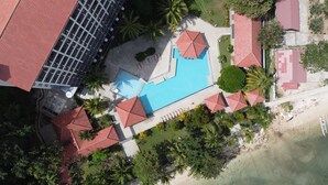An aerial view of a resort with a swimming pool, palm trees, and red-roofed buildings. - LAPRIMA Hotel Flores (Labuan Bajo)