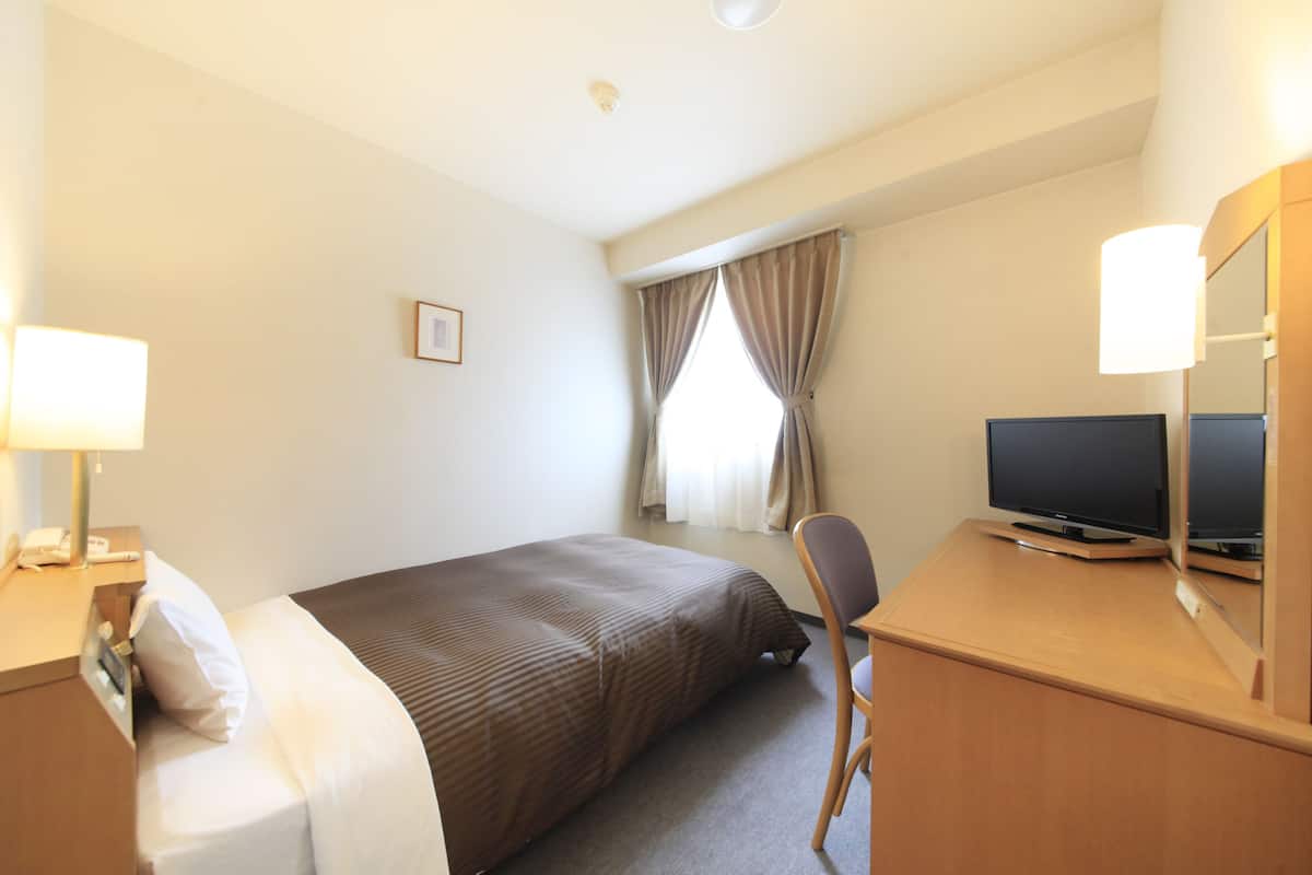 Une chambre d’hôtel avec un lit, un bureau avec une télévision, une chaise, une lampe et une fenêtre avec des rideaux.