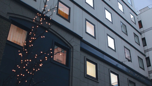A multi-story hotel building with a modern architectural design, featuring a series of uniformly sized windows and a visible entrance with a glass door. - Hotel Morschein (Matsumoto)