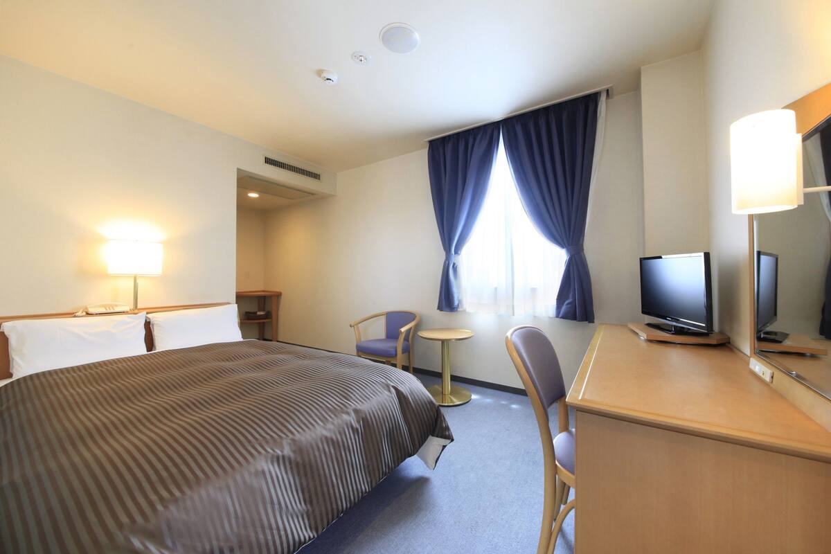 Une chambre d’hôtel avec un grand lit, un bureau avec une télévision, une chaise, une petite table ronde et une fenêtre avec des rideaux.