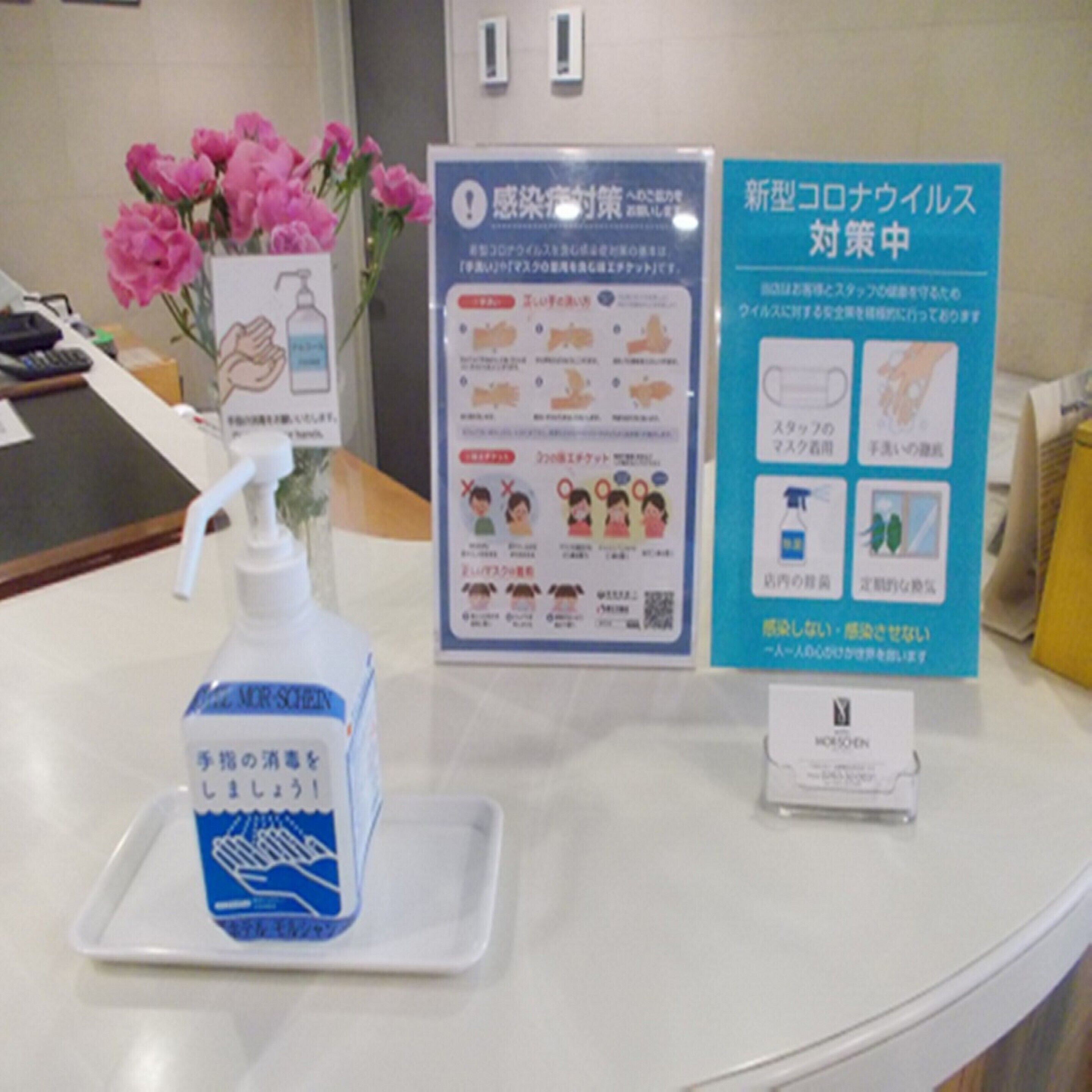 Un mostrador de recepción con un dispensador de gel hidroalcohólico, un jarrón de flores rosadas y carteles informativos.