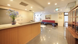A reception area with a wooden counter, a vase of flowers, a seating area with a red couch, and a glass cabinet. - Hotel Morschein (Matsumoto)