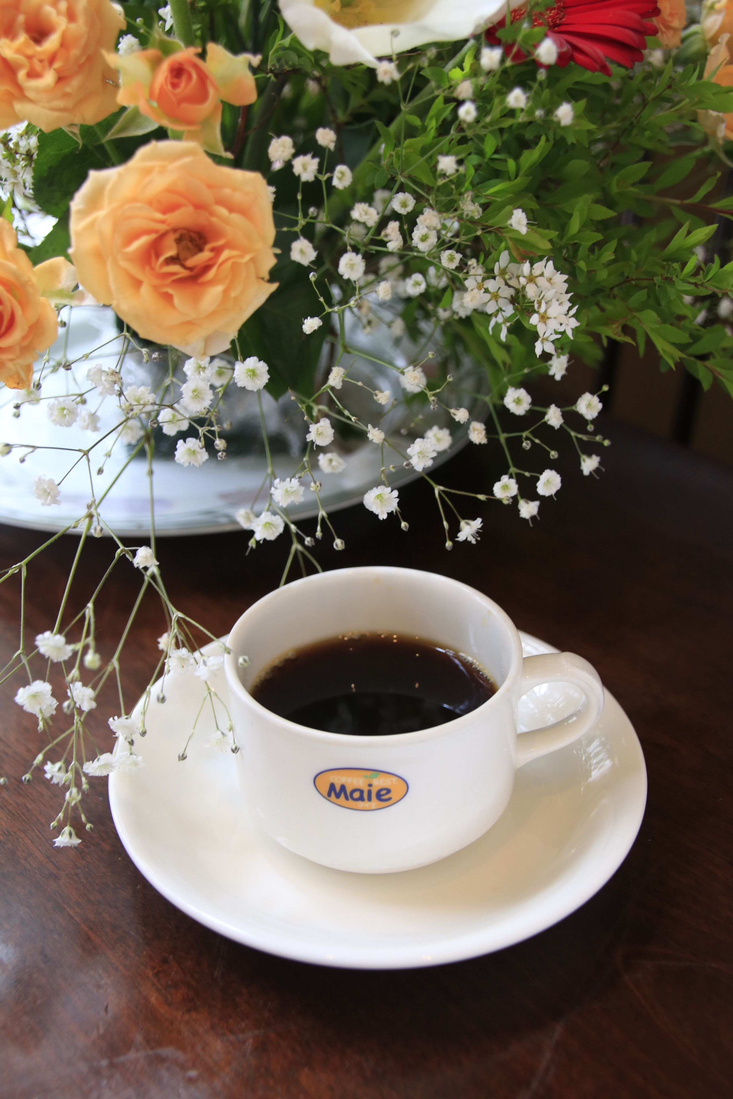 A cup of coffee on a saucer with a logo, placed on a table with a floral arrangement in the background.