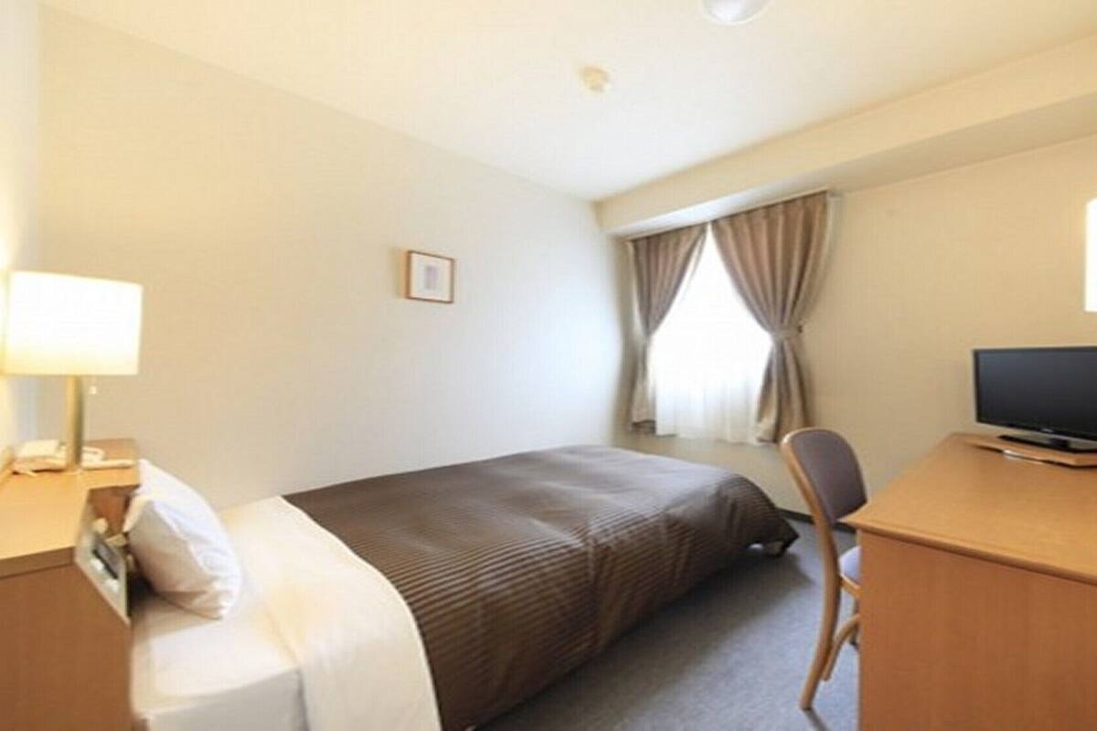 Une chambre d’hôtel avec un lit, un bureau avec une télévision, une chaise, une lampe et une fenêtre avec des rideaux.