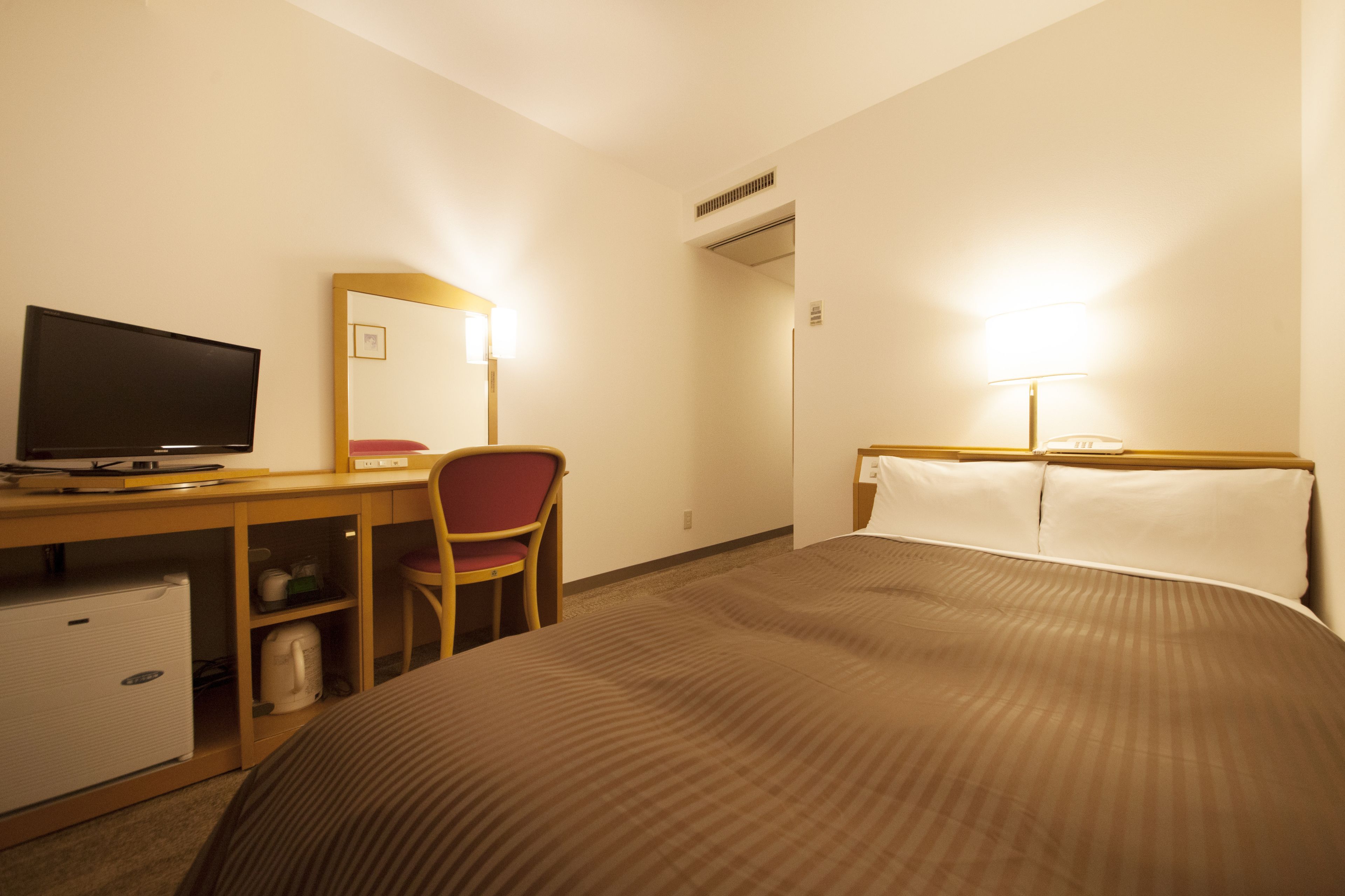 A hotel room with a bed, a desk with a computer, a chair, and a small refrigerator.