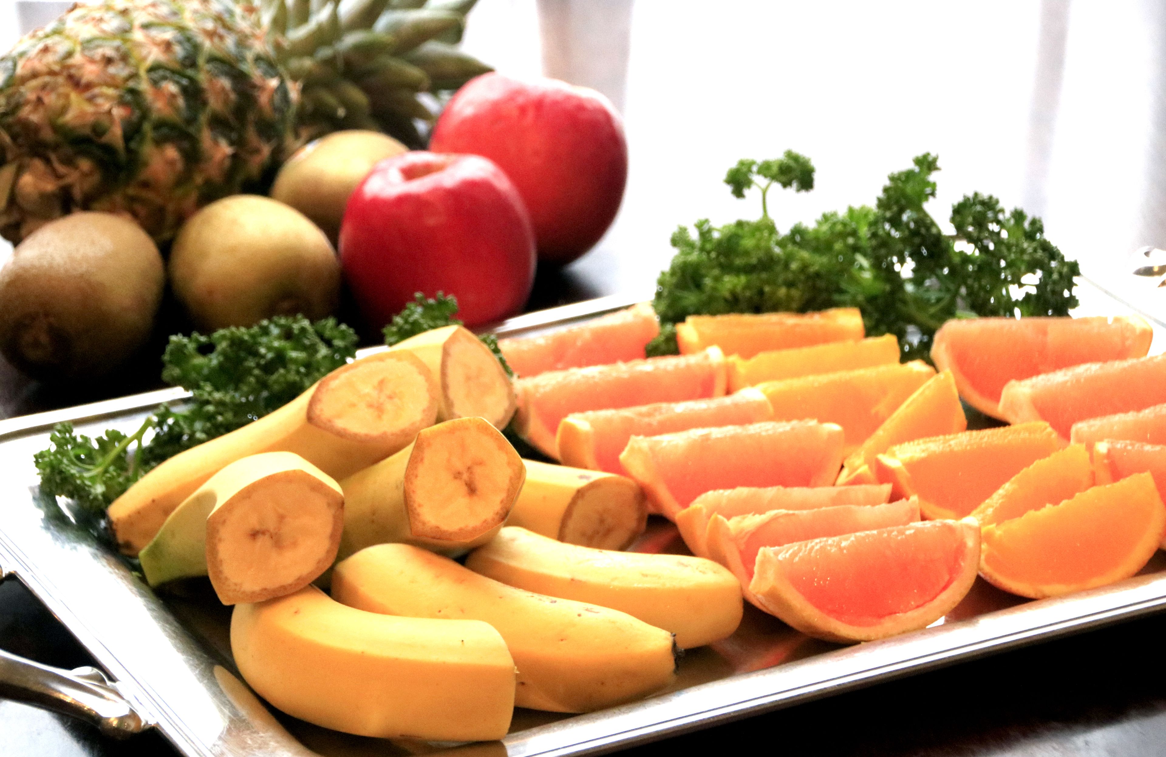 Una bandeja con frutas cortadas, como plátanos, naranjas y manzanas, con una piña y kiwi al fondo.