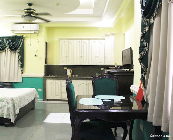 A compact hotel room with a bed, a dining table with chairs, a small kitchen area, and a ceiling fan.