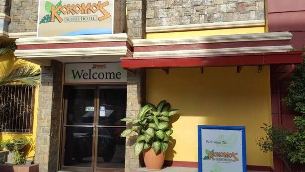 Entrada de un hotel con un letrero de 'Welcome', una planta grande en maceta y un cartel de bienvenida enmarcado.