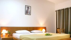 A bedroom with a wooden bed, white bedding, two bedside lamps, a nightstand, a framed picture on the wall, and a window with vertical blinds. - Dream Valley Resort (Hyderabad)