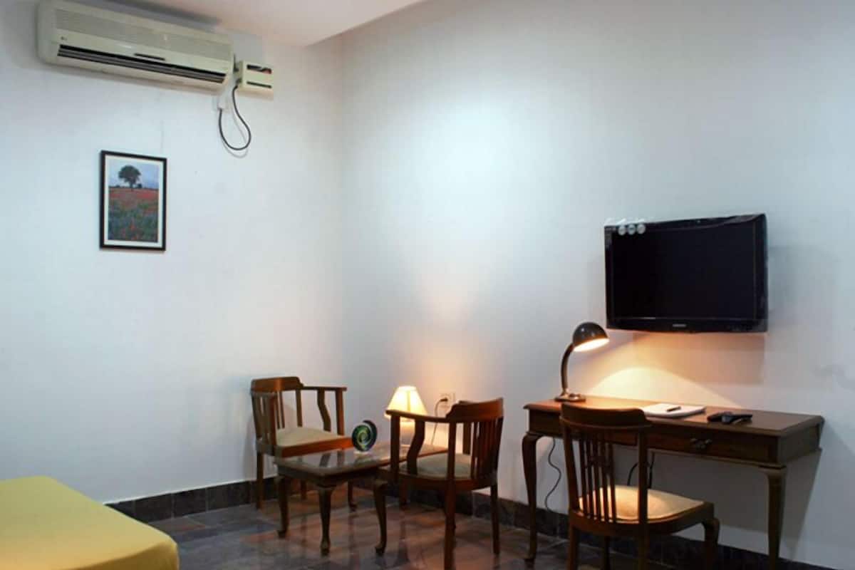 Una habitación pequeña y ordenada con escritorio, dos sillas, cama, televisión y aire acondicionado.