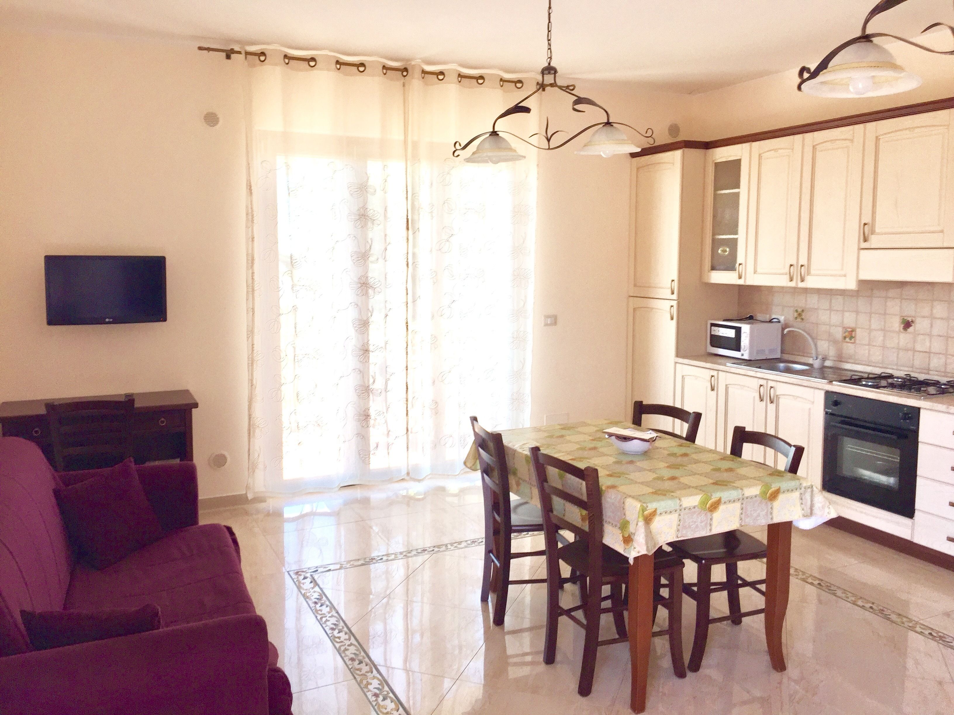 Una sala de estar con sofá y televisor, un comedor con mesa y sillas, y una cocina con armarios y electrodomésticos.