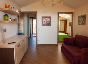 A living room with a wooden floor, a red sofa, a kitchen area with wooden cabinets, and a hallway leading to another room.