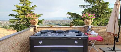 A hot tub with a view of a cityscape, surrounded by potted plants and a table with a red tablecloth.