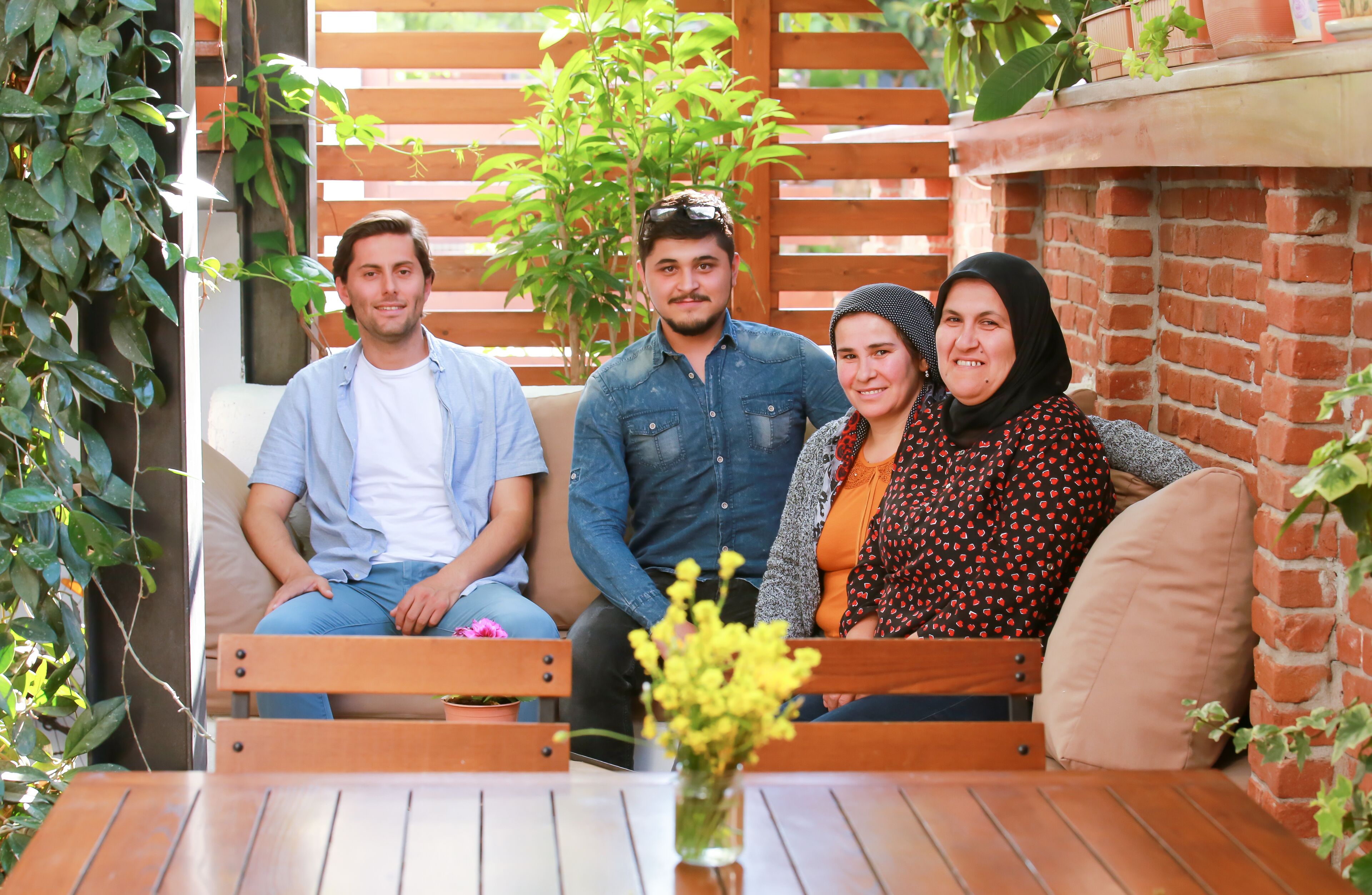 A group of people seated on a wooden bench in a covered outdoor area with brick walls and wooden slats.