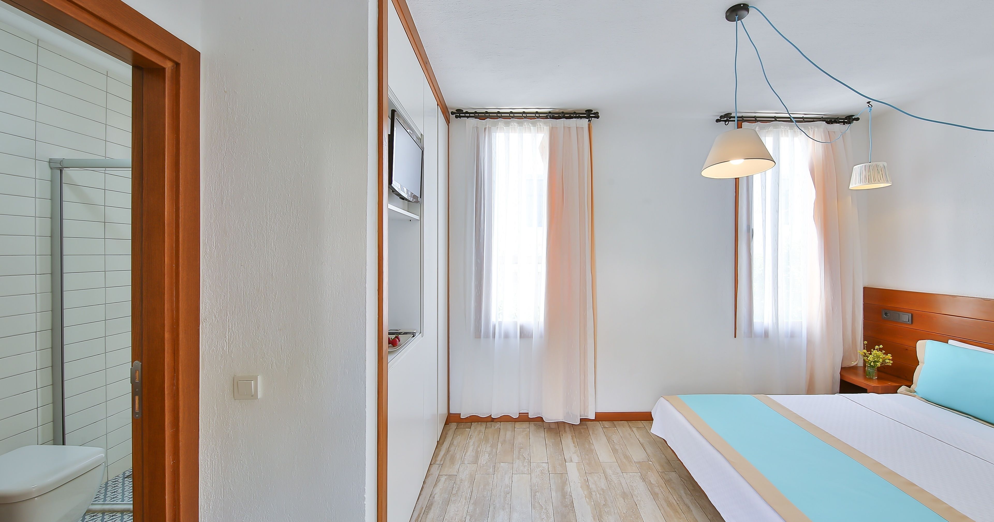 Una habitación de hotel con cama, mesita de noche, ventana con cortinas, televisor montado en la pared y puerta de madera.