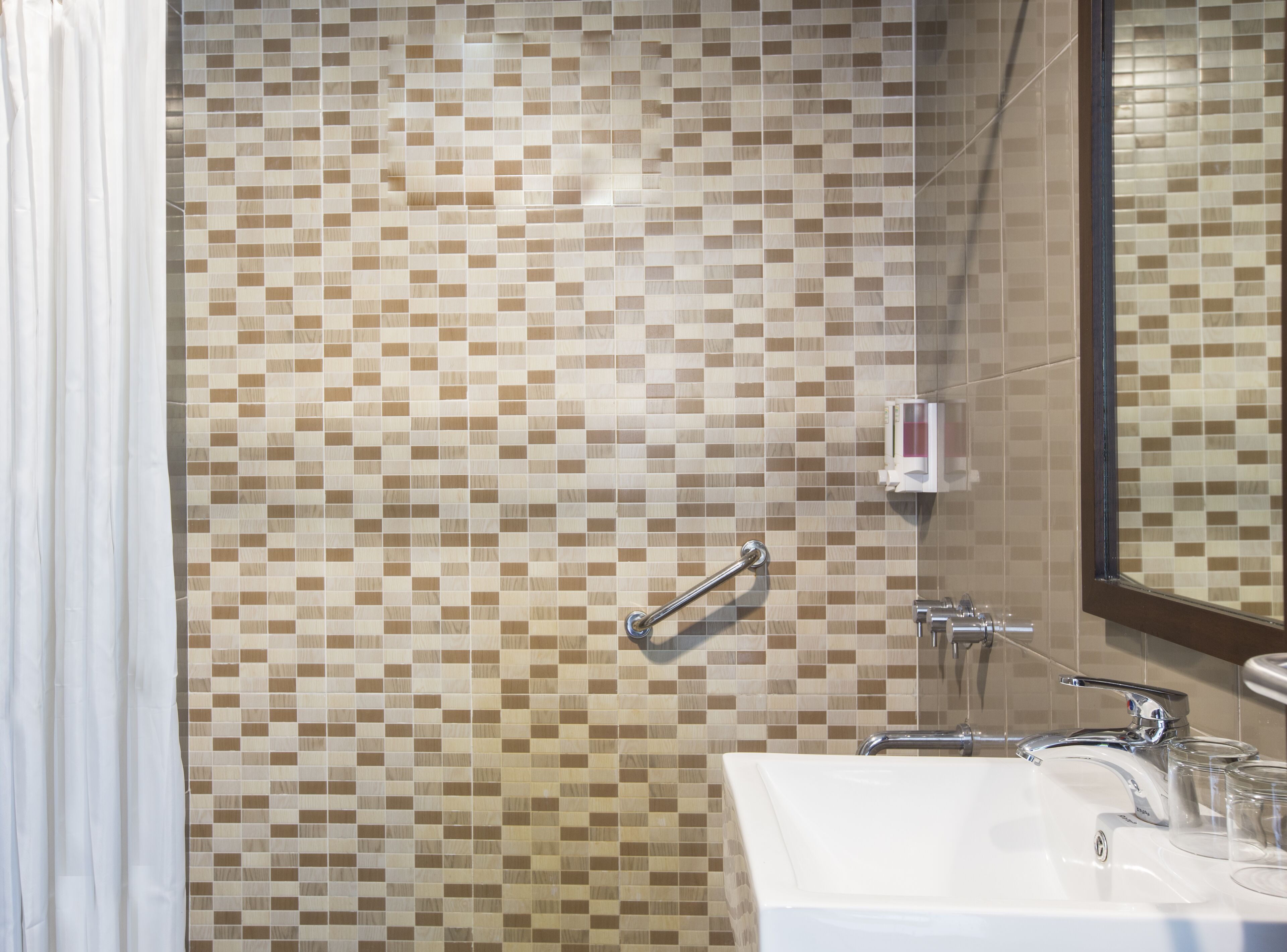Un baño con pared de mosaicos, ducha con barra de apoyo, lavabo con grifo y espejo.