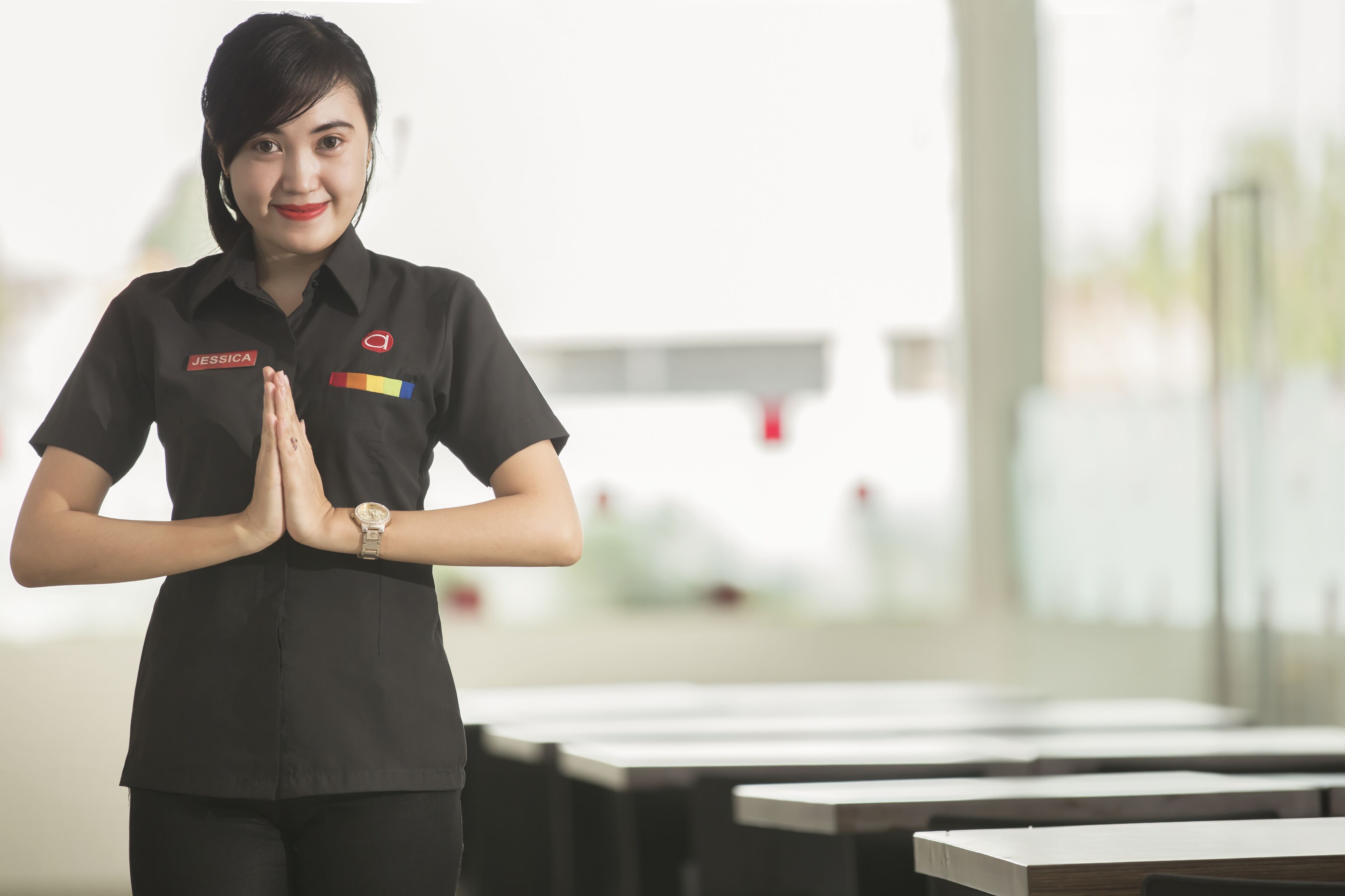 Une personne vêtue d’un uniforme noir, portant une étiquette avec son nom et une montre, se tient dans une pièce où se trouvent des tables et des chaises.
