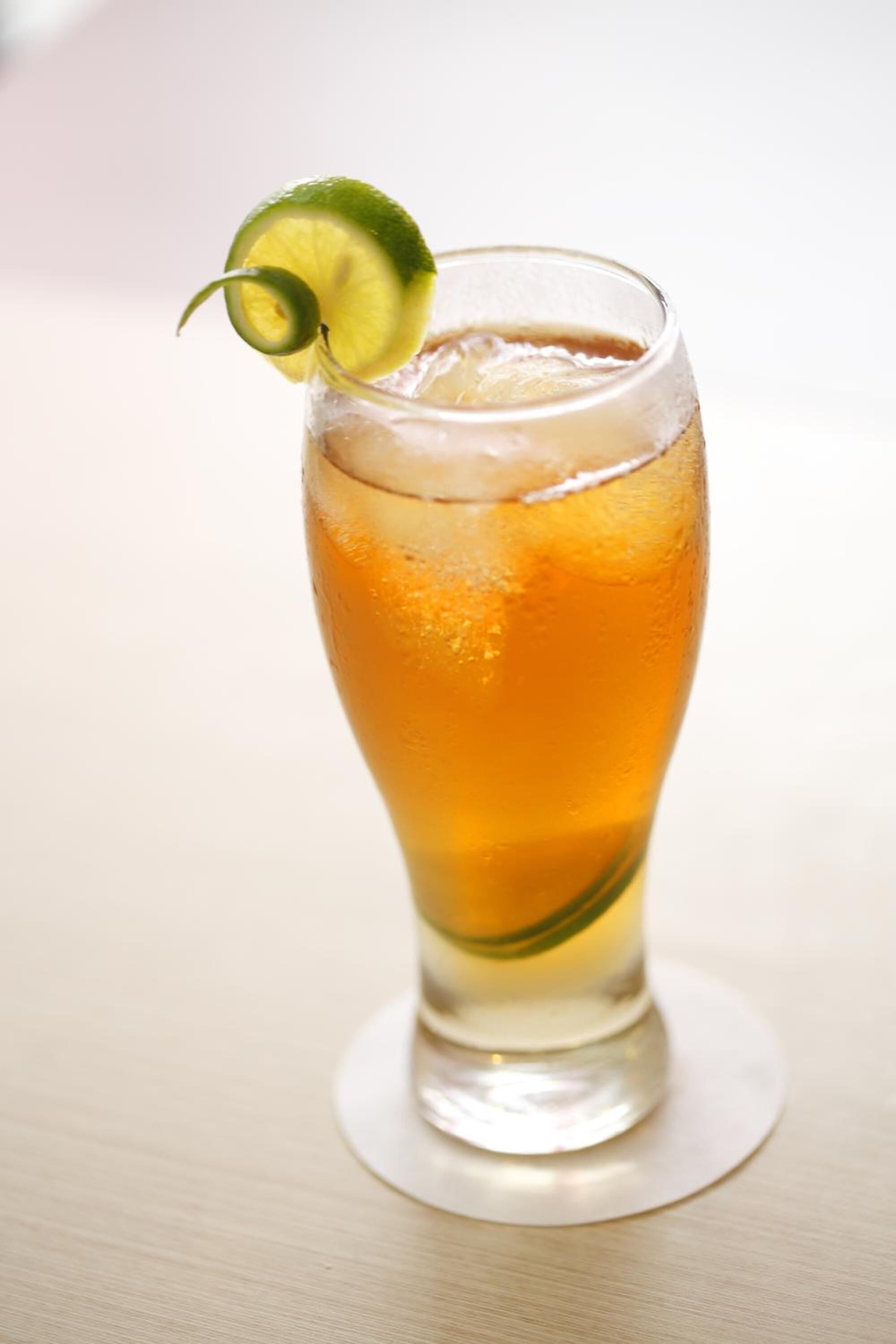 Un grand verre contenant un liquide clair, peut-être un cocktail, agrémenté d’une tranche de citron vert et posé sur un sous-verre rond.