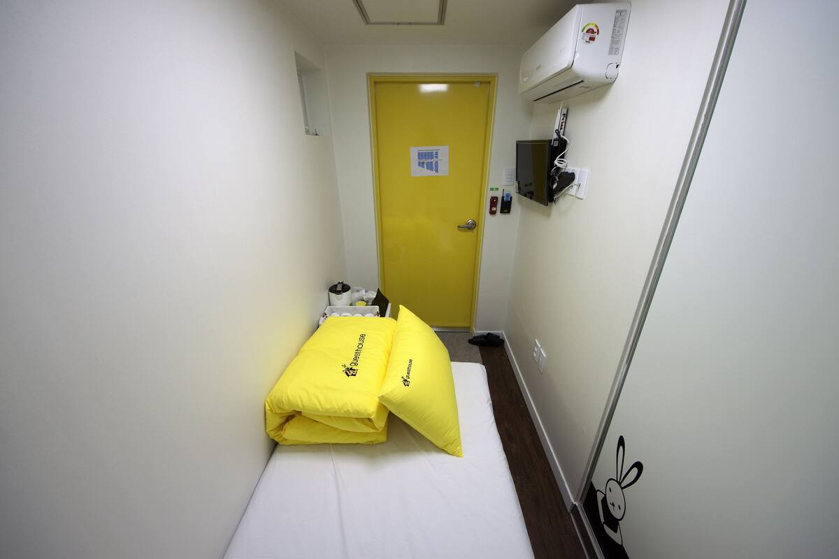 A narrow hotel room with a bed, a yellow door, and a wall-mounted air conditioning unit.